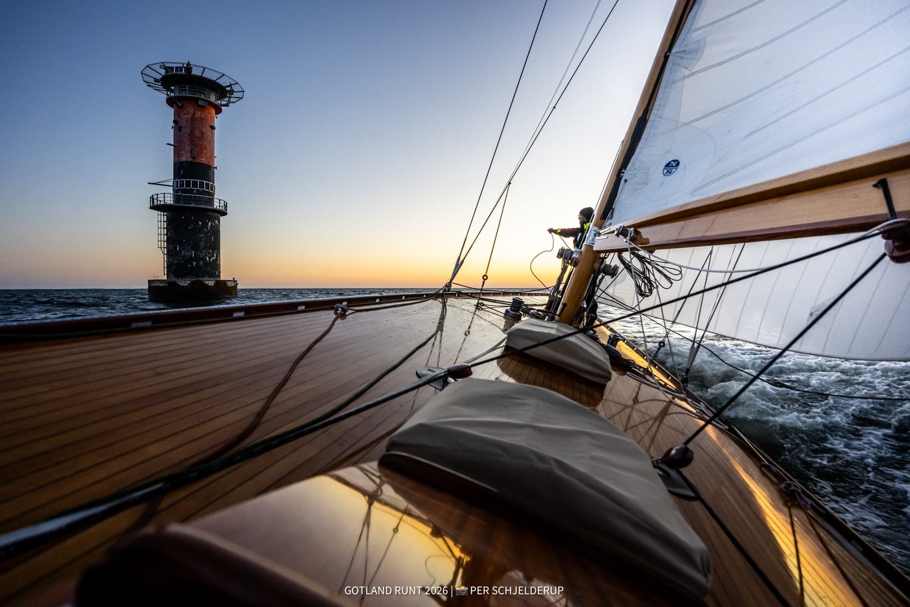 The foredeck of a wooden sailing boat at sunset, a lighthouse off the bow.